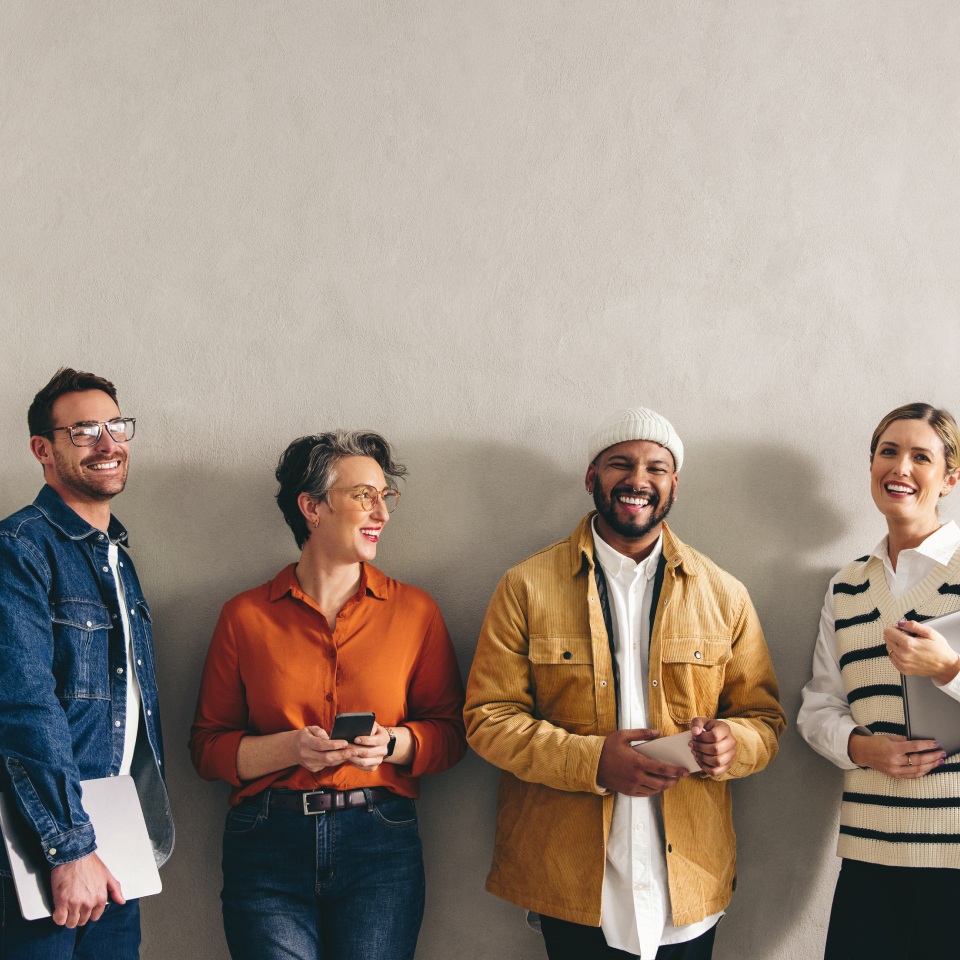 A group of four smiling people stand against a plain wall. From left to right: a man in glasses holding a laptop, a person with short gray hair holding a phone, a man wearing a beanie and a yellow jacket, and a woman in a white sweater with black stripes.