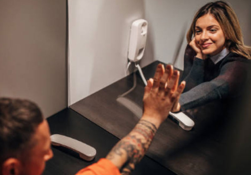 A woman and an incarcerated man are separated by a glass partition in a visiting area. The woman, smiling with her chin resting on her hand, mirrors a high-five gesture with the man. A phone handset is visible on the counter next to them.