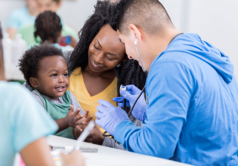 A healthcare worker in blue scrubs and gloves uses a stethoscope on a smiling toddler held by a woman in a yellow shirt. The scene takes place in a medical setting with other people in the background.