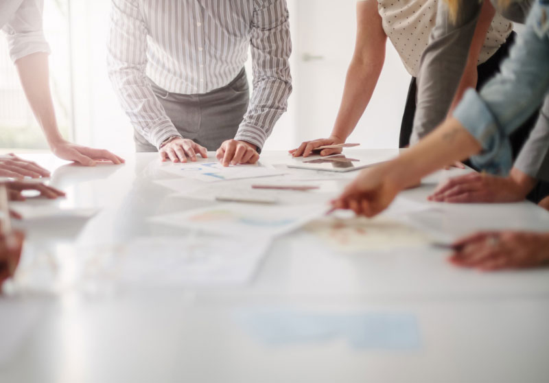 A group of people in business attire lean over a white table collaborating on a project. Papers, pens, and a tablet are spread out on the table, and their hands are actively engaged in discussion and pointing to various items. The setting suggests a team meeting.