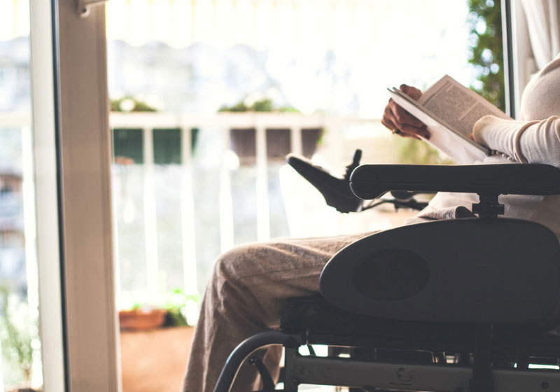 A person sitting in a wheelchair by a window, reading a book. Sunlight is streaming in, creating a warm and cozy atmosphere. The person is wearing light-colored, comfortable clothes, and there are plants visible on the balcony outside.