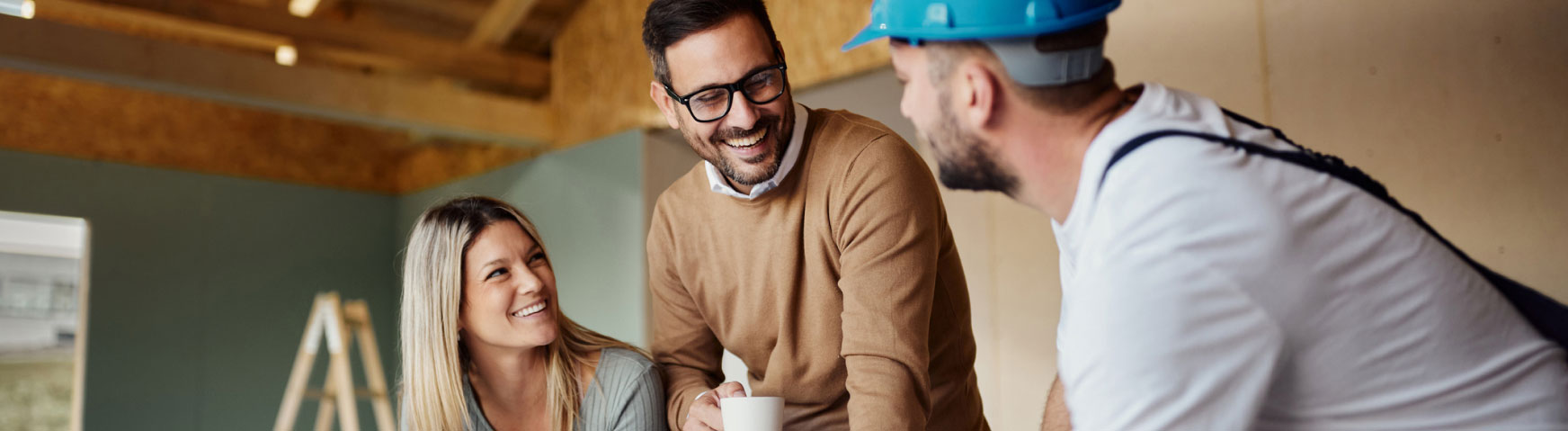 A woman and two men are engaged in conversation at a construction site. One man is holding a cup and smiling, while another man wearing a blue hard hat appears to be explaining something to them. A ladder is visible in the background.