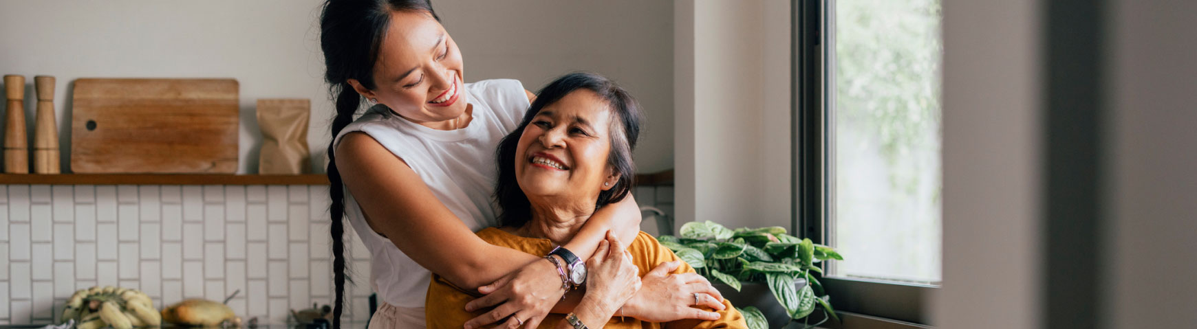 A young woman embraces an older woman from behind while they both smile in a kitchen. The kitchen has a modern, minimalist design with white tiles, wooden cutting boards, and various potted plants visible in the background. Light streams in from a nearby window.