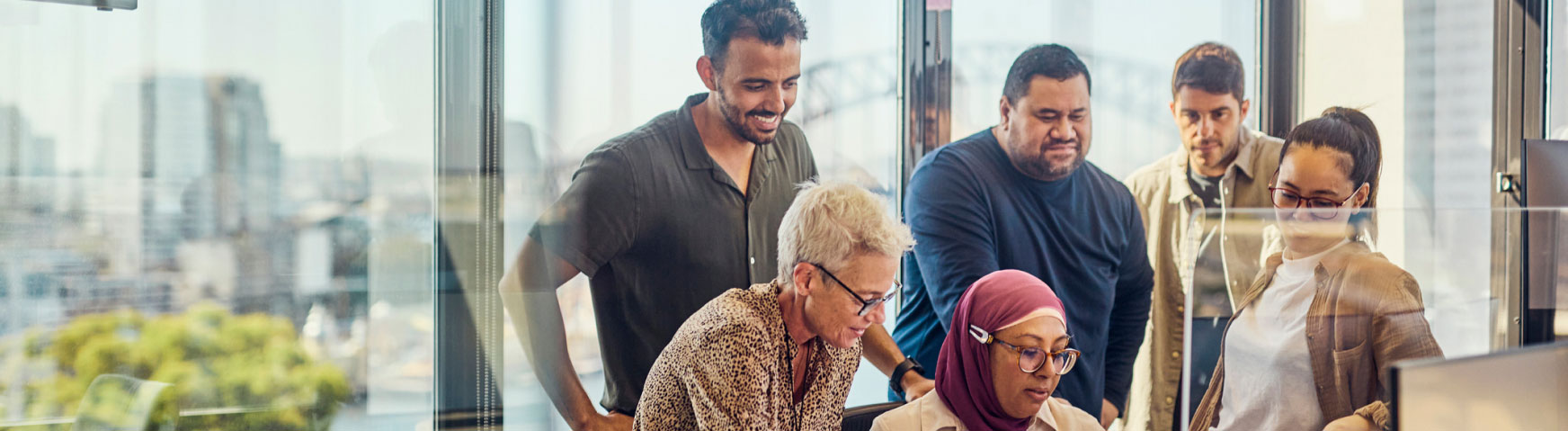 A diverse group of five colleagues gathers around a computer, engaged in discussion in a bright office with city views visible through the large windows in the background. One person is typing while the others are watching attentively.