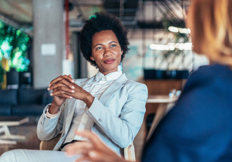 A person is sitting in an office environment, wearing a light gray suit and white shirt. They have short curly hair and are smiling slightly while clasping their hands together. Another person, slightly out of focus, is seated and facing them.