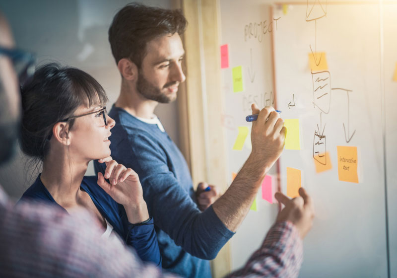 Two people collaborate on a project, writing and drawing on a whiteboard covered with colorful sticky notes and diagrams. One person holds a marker and explains while the other observes and listens attentively. The setting appears to be an office or meeting room.