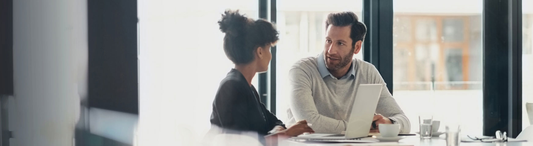 A woman and a man are seated at a table in an office setting, having a discussion. The man is looking at the woman while holding a laptop. They appear engaged in conversation, with large windows in the background allowing natural light to fill the room.