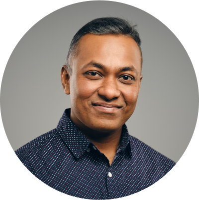 A man with short hair smiles gently at the camera. He is wearing a dark, patterned button-up shirt and is captured against a simple gray background. His expression is relaxed and content. The photo is cropped in a circular frame.