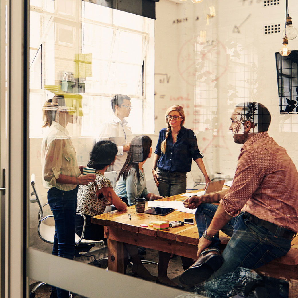 A group of six people are having a discussion around a conference table in a modern office, with large windows and notes on a transparent wall. Some are seated while others stand, actively engaging in conversation. Office supplies are scattered on the table.