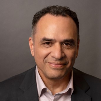 The image shows a headshot of Luis Garcia with short dark hair, smiling without teeth. They are wearing a black suit with light collared shirt.