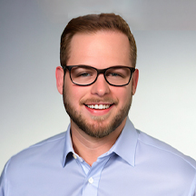 Timothy Gentile, a man wearing a blue dress shirt and black-rimmed glasses smiles at the camera.