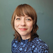 Joanna Prout, a woman with short, light brown hair is smiling at the camera. She is wearing a blue top with a floral pattern. The background is a plain blue-gray color.