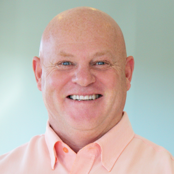 Charles is smiling at the camera. He is wearing a light pink collared shirt and is set against a plain, softly-lit background.